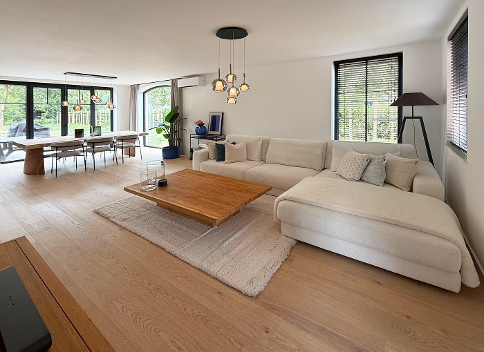 Ruime moderne woonkamer met licht houten vloer, een grote witte bank, houten salontafel, eethoek met een lange tafel en stoelen, grote ramen en kamerplanten. Natuurlijk licht vult de ruimte.