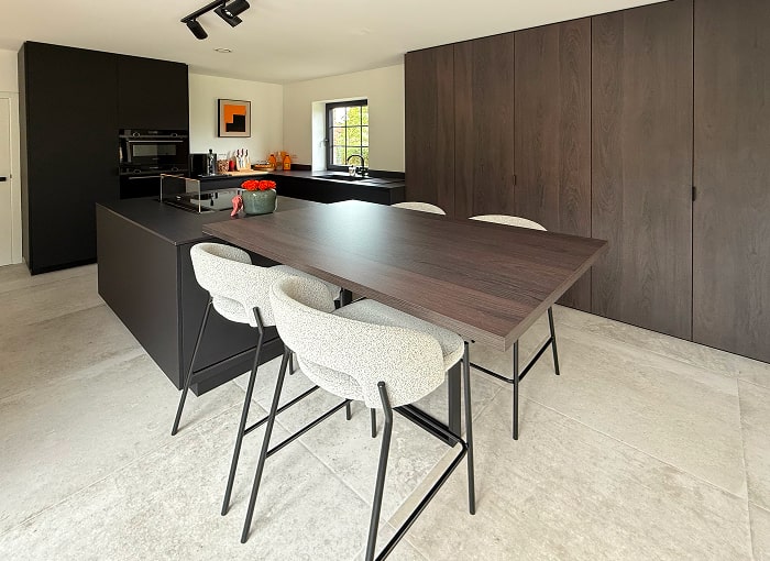 Moderne keuken met donkerhouten kasten, een groot eiland en een hoge eettafel met drie gestoffeerde stoelen. Lichtgrijze tegelvloeren en een raam dat natuurlijk licht binnenlaat maken het minimalistische ontwerp compleet.