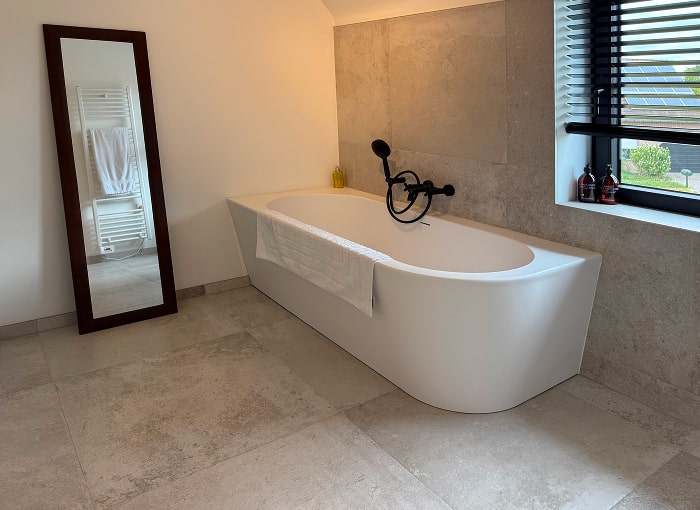 Moderne badkamer met een wit vrijstaand bad, een handdoek die over de rand hangt, een grote spiegel op de vloer, grijs betegelde muren en vloeren en een raam met zwarte jaloezieën die daglicht binnenlaten.