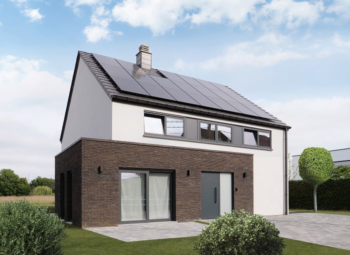 Une maison moderne à deux étages avec des briques sombres et des murs blancs, de grandes fenêtres et des panneaux solaires sur le toit, entourée d'une pelouse verte, de buissons et d'un petit arbre, sous un ciel bleu avec quelques nuages.
