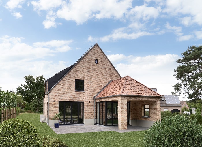 Een modern bakstenen huis met grote ramen, een schuin dak en een overdekte patio ligt op een groen gazon onder een gedeeltelijk bewolkte hemel. Op de achtergrond zijn zonnepanelen zichtbaar op een naburig huis.
