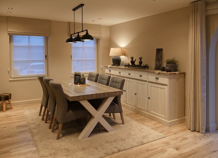 Een gezellige eetkamer met een houten tafel, acht stoelen met kussens, een beige vloerkleed, zwarte hanglampen en een dressoir versierd met lampen en ornamenten, alles in warme, neutrale tinten.