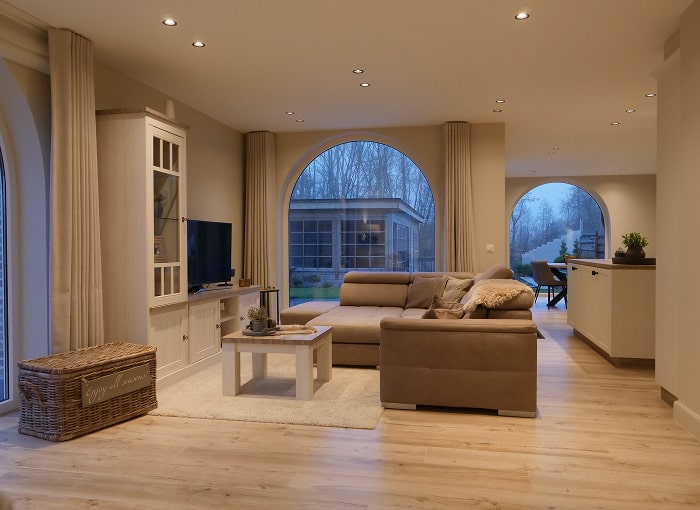 Een gezellige woonkamer met een bruine slaapbank, lichthouten vloeren, een tv op een witte kast en grote boogramen die uitzicht bieden op een tuin en bomen buiten. Aan de linkerkant van de kamer staat een rieten mand.