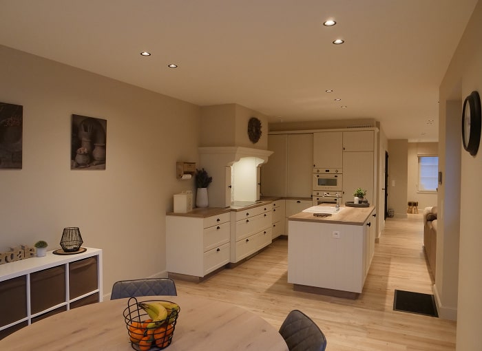 Een moderne keuken en eethoek met lichthouten vloeren, crèmekleurige kasten, een eiland, ingebouwde plafondverlichting en een ronde eettafel met stoelen en een fruitschaal op de voorgrond.