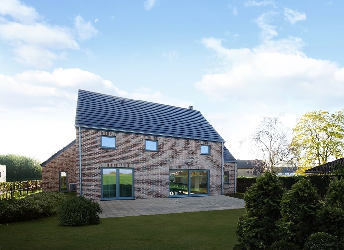 Une maison moderne en briques avec un toit en tuiles sombres, de grandes fenêtres et un patio spacieux se trouve dans une cour verte sous un ciel bleu avec des nuages épars. Des arbustes et des arbres entourent la maison.