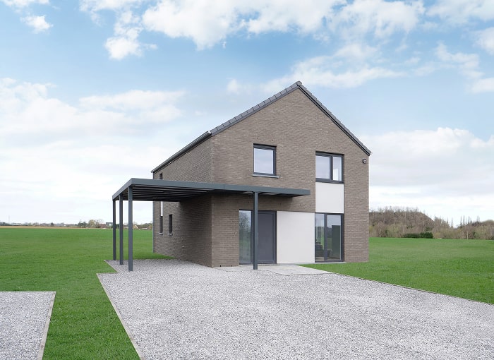 Een modern huis van twee verdiepingen met grijze bakstenen muren, grote ramen en een carport met plat dak staat op een oprit van grind, omgeven door een groen gazon onder een gedeeltelijk bewolkte hemel.