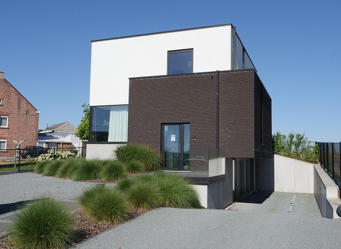 Maison moderne à deux étages avec un étage supérieur blanc et un étage inférieur en briques foncées, de grandes fenêtres et une allée en pente entourée de graminées ornementales sous un ciel bleu clair.