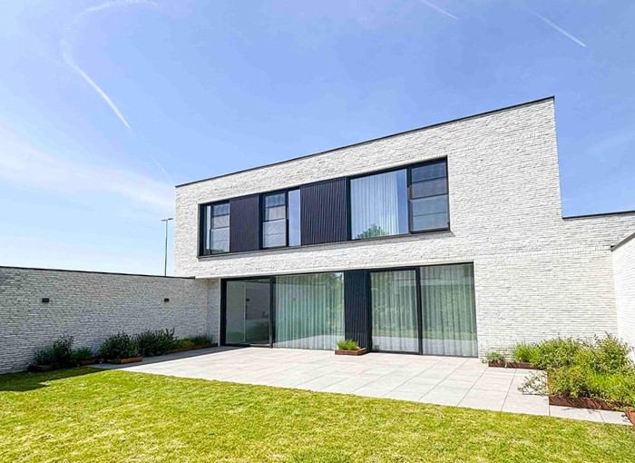Une maison moderne à deux étages avec un toit plat, de grandes fenêtres, un extérieur en briques blanches et un patio spacieux. La maison fait face à une pelouse verte sous un ciel bleu clair.