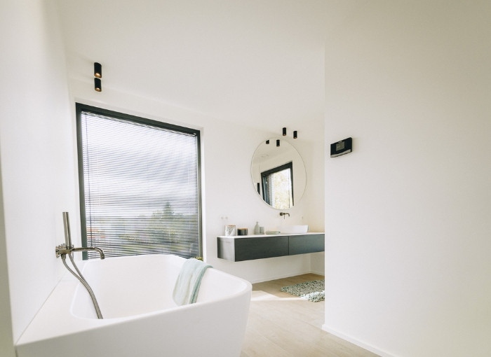 Une salle de bains moderne et minimaliste avec une baignoire blanche près d'une grande fenêtre avec des stores, un miroir rond au-dessus d'un lavabo flottant et un sol en bois clair. Une serviette est suspendue au bord de la baignoire.