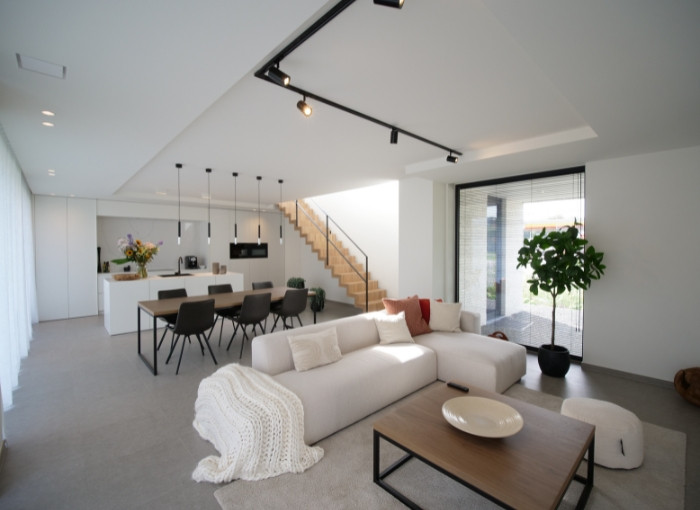 Un salon et une salle à manger modernes et ouverts, avec un canapé sectionnel blanc, une table basse en bois, une table à manger avec des chaises noires, un escalier, de grandes fenêtres et un décor minimaliste.
