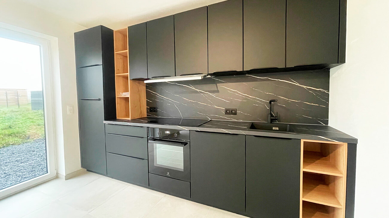 Cuisine moderne avec placards noirs mats, dosseret en marbre noir, four encastré et étagères ouvertes en bois. Il y a un évier sur la droite et une grande fenêtre sur la gauche qui donne sur une cour herbeuse.