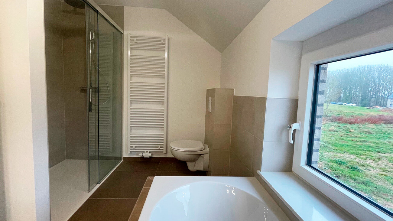 Salle de bains moderne avec une douche en verre, des toilettes murales, un porte-serviettes chauffant et une baignoire à côté d'une grande fenêtre donnant sur un paysage extérieur herbeux. Le design est minimal et lumineux.