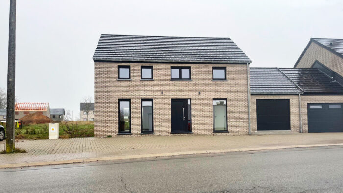 Une maison moderne en briques à deux étages avec un toit en tuiles sombres, de grandes fenêtres et un garage attenant, située dans une allée pavée le long d'une rue tranquille par un temps couvert.