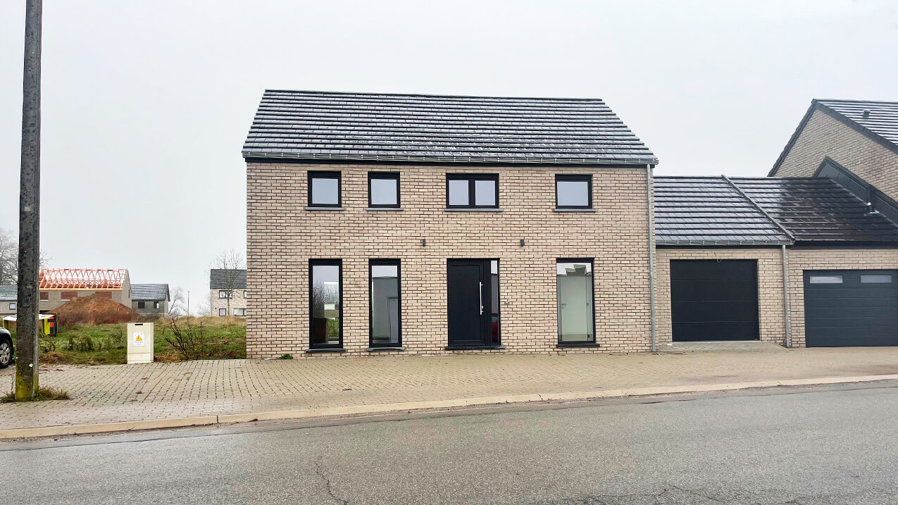 Une maison moderne en briques à deux étages avec un toit en tuiles sombres, de grandes fenêtres et un garage attenant, située dans une allée pavée le long d'une rue tranquille par un temps couvert.