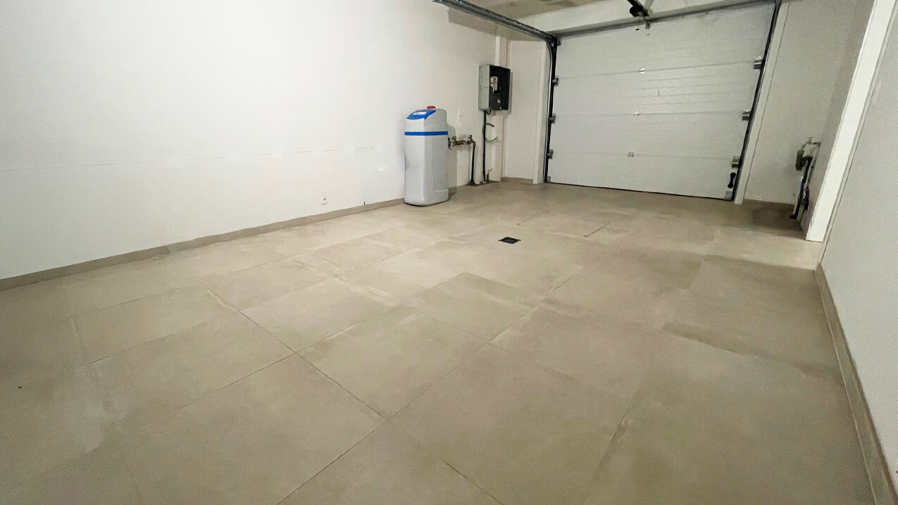 Un garage vide et propre avec un sol carrelé de couleur claire, des murs blancs, une porte de garage fermée, un réservoir d'adoucisseur d'eau et des panneaux utilitaires le long du mur du fond.
