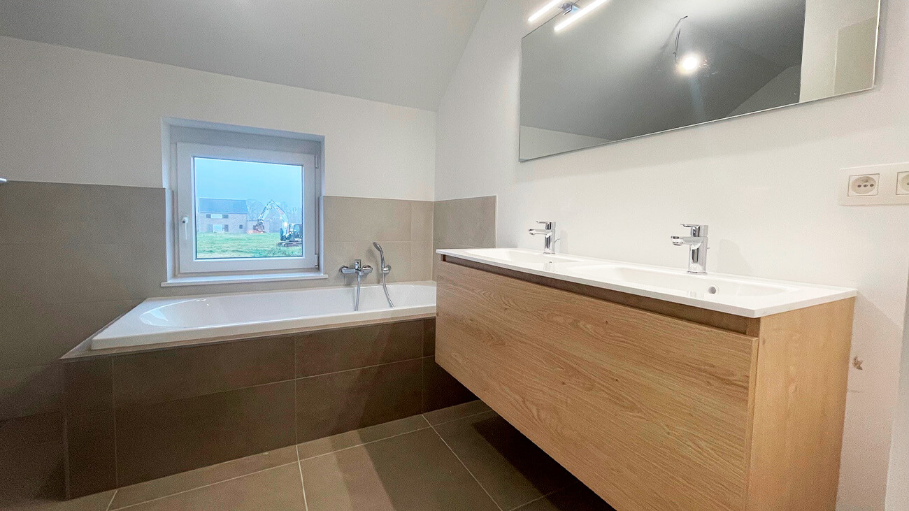 Salle de bains moderne avec une grande baignoire sous une fenêtre, un meuble à double vasque avec des armoires en bois, un grand miroir au-dessus, et des carreaux gris clair sur le sol et les murs.