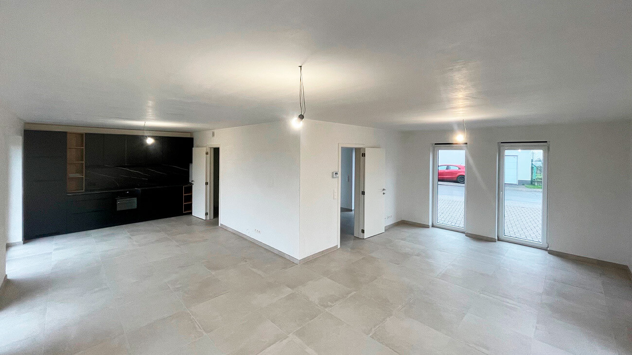 Pièce spacieuse, moderne, non meublée, à aire ouverte, avec sol carrelé, murs blancs, deux ampoules suspendues, un coin cuisine noir au fond à gauche, et de grandes fenêtres et portes menant à l'extérieur.