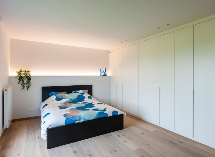Chambre à coucher minimaliste avec un lit double à la literie bleue et blanche, des armoires blanches encastrées le long d'un mur, un sol en bois clair, un radiateur, une plante en pot et des objets de décoration bleus sur une étagère avec un éclairage tamisé.