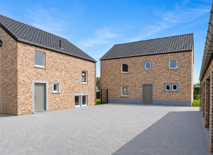 Une cour pavée entourée de trois maisons modernes en briques aux toits sombres et aux portes et fenêtres grises, sous un ciel d'un bleu éclatant parsemé de quelques nuages légers.