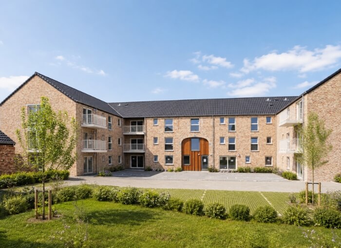Un immeuble d'appartements moderne de trois étages en briques, doté de balcons et de grandes portes centrales en bois, entouré de pelouses verdoyantes, de jeunes arbres et d'arbustes, sous un ciel bleu parsemé de nuages.