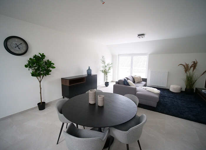 Salon et salle à manger modernes avec une table ronde noire, quatre chaises grises, un canapé sectionnel gris, des plantes, une horloge murale et de grandes fenêtres laissant entrer la lumière naturelle. La décoration est minimale et contemporaine.