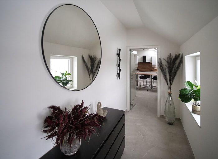 Un couloir moderne et minimaliste avec des murs blancs, un miroir rond au-dessus d'une commode noire, des plantes décoratives et un aperçu du coin repas avec une table et des chaises. L'espace est baigné de lumière naturelle.