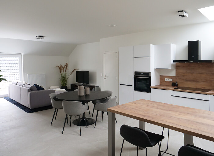 Cuisine et salon modernes à aire ouverte avec des armoires blanches, des comptoirs en bois, une table à manger ronde avec des chaises grises, un canapé sectionnel gris et une télévision. L'espace est lumineux, avec des couleurs neutres et un décor minimaliste.