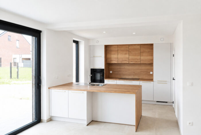 Cuisine moderne avec des éléments blancs, des plans de travail en bois, un four encastré, un évier et une grande fenêtre. La lumière naturelle illumine cet espace minimaliste, et la porte vitrée offre une vue sur l'extérieur.