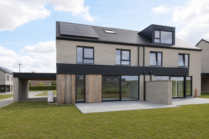 Maison moderne à deux étages, avec une façade en briques beiges, un toit noir, de grandes fenêtres et des lambris en bois. Elle dispose d'un petit patio et d'une grande pelouse verte à l'avant. Le ciel est partiellement nuageux.