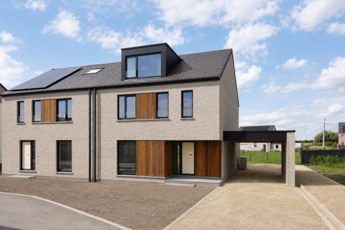 Maison jumelée moderne à deux étages, avec une façade en briques grises, des éléments décoratifs en bois, de grandes fenêtres et une allée sous un abri pour voiture, se découpant sur un ciel partiellement nuageux.
