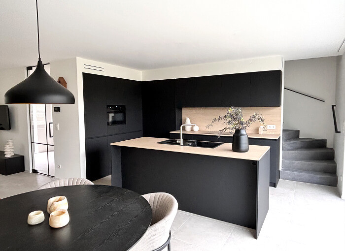 Moderne keuken met matzwarte kasten en een licht houten aanrechtblad op het eiland. Een zwarte hanglamp hangt boven een ronde eettafel met lichte stoelen. De trap is zichtbaar aan de rechterkant en de ruimte is licht en minimalistisch.