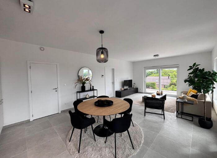 Salon moderne et ouvert, avec une table à manger ronde, des chaises noires, un canapé, une télévision, une grande fenêtre, une plante en pot et une décoration minimaliste. Une lumière naturelle abondante inonde la pièce.