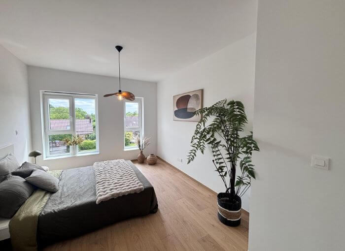 Chambre à coucher minimaliste comprenant un lit, une literie aux tons neutres, de grandes fenêtres, un plafonnier, des plantes en pot, un parquet et des œuvres d'art abstraites accrochées aux murs. La lumière naturelle inonde la pièce, créant une atmosphère lumineuse et apaisante.
