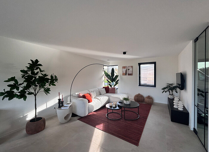Een moderne woonkamer met een witte bank, rode kussens, een rood vloerkleed, twee ronde salontafels, grote kamerplanten en decor. De kamer heeft witte muren, grote ramen en veel natuurlijk licht.