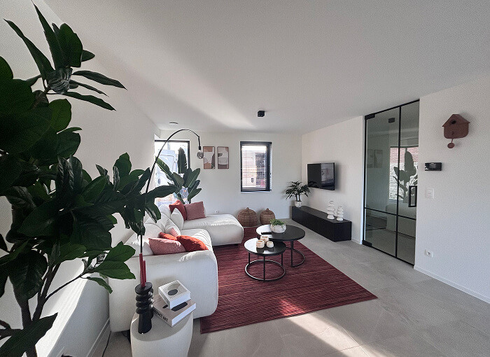 Een lichte, moderne woonkamer met witte muren en grote ramen. Er staat een witte L-vormige bank met rode kussens, een rood vloerkleed, ronde salontafels, groene planten en een tv aan de muur tegenover de zithoek.