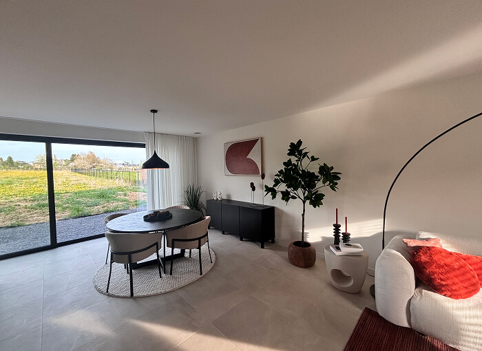 Een moderne woon- en eetruimte met grote ramen die uitkijken op een veld, een ronde eettafel met vier stoelen, een witte bank, een plant in potten, een dressoir, abstracte muurkunst en een neutrale inrichting met rode accenten.