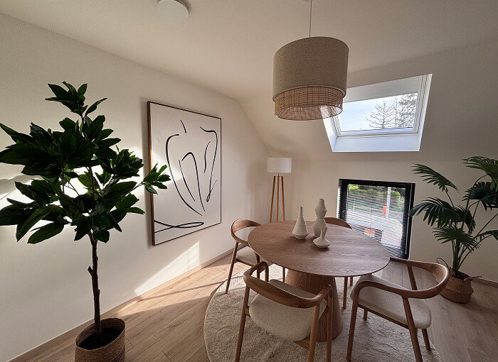 Een moderne eetkamer met een ronde houten tafel, vier stoelen, abstracte muurkunst, potplanten, een groot raam en een plafondlamp, allemaal verlicht door natuurlijk zonlicht dat naar binnen stroomt.