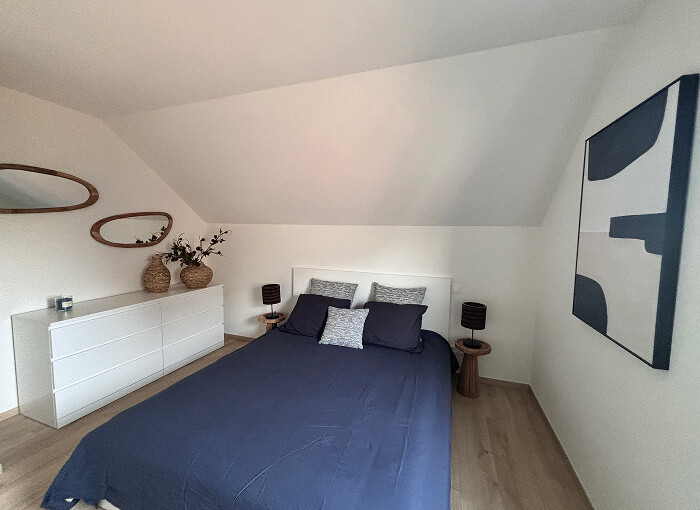 Een moderne slaapkamer met een schuin plafond, blauw beddengoed, een wit dressoir met vazen en decor, ovale spiegels aan de muur, twee kleine nachtkastjes met lampen en een groot abstract kunstwerk.