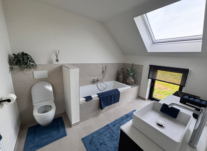 Moderne badkamer met een bad, toilet en twee wastafels. Blauwe handdoeken en matten accentueren de lichtgekleurde ruimte. Een groot dakraam en raam laten natuurlijk licht binnen. Een plant en decor aan de muur achter het toilet.