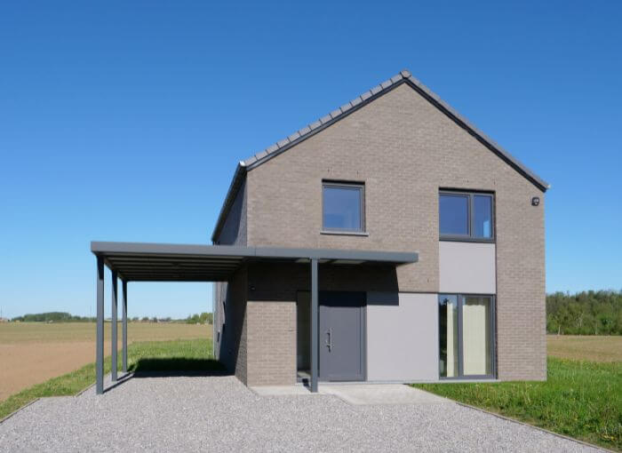 Maison moderne à deux étages, avec une façade en briques grises, de grandes fenêtres et un abri pour voiture attenant, située au bout d'une allée en gravier, avec des champs et un ciel bleu limpide en arrière-plan.