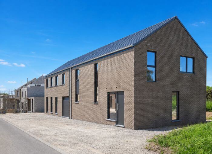 Maison moderne en briques à deux étages, dotée de grandes fenêtres, d'un toit en pente de couleur foncée et d'une allée en gravier, sous un ciel bleu limpide. D'autres maisons en cours de construction sont visibles à l'arrière-plan.