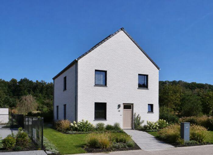 Une maison moderne de deux étages, en briques blanches, avec un toit gris, des fenêtres aux encadrements noirs et une porte marron clair. La maison est entourée d'un espace vert et se détache sur un ciel d'un bleu limpide.