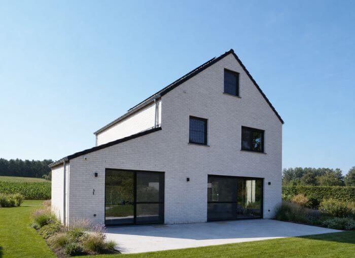 Maison moderne à deux étages en briques blanches, dotée de grandes fenêtres aux encadrements noirs et d'un toit en pente, entourée de pelouse, de plantes et de champs à perte de vue sous un ciel bleu limpide.
