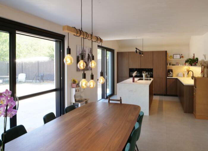 Cuisine et coin repas modernes avec une table en bois, des chaises vertes, des suspensions, de grandes fenêtres et des placards marron. À l'arrière-plan, un îlot de cuisine avec un plan de travail blanc. Ambiance lumineuse et accueillante.