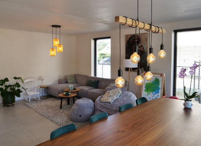 Salon moderne avec une table à manger en bois, des ampoules suspendues, un canapé modulable gris, des tables basses, des poufs, des plantes et de grandes fenêtres laissant entrer la lumière naturelle.