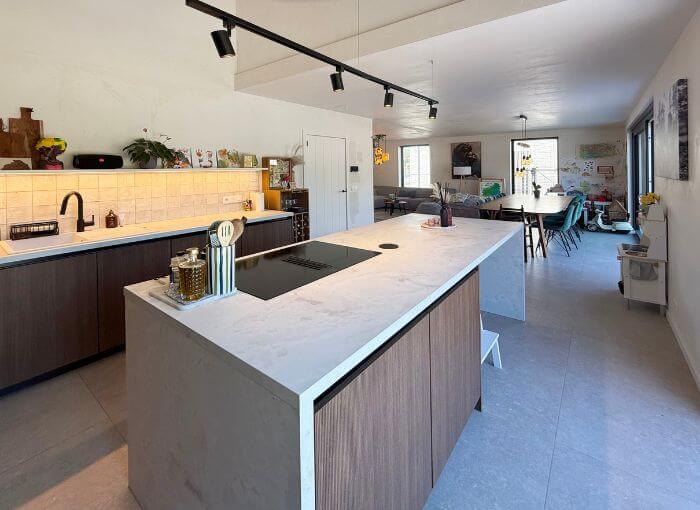 Cuisine moderne dotée d'un grand îlot en marbre, d'une cuisinière encastrée, d'armoires en bois, de robinetterie noire et d'un agencement ouvert donnant sur un espace salon-salle à manger lumineux. La décoration comprend des fleurs, des bocaux et des œuvres d'art murales.