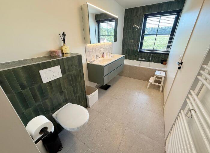 Salle de bains moderne avec un mur carrelé de vert, un meuble-lavabo suspendu avec miroir, une baignoire près de la fenêtre, des toilettes suspendues, un tabouret et un radiateur sèche-serviettes. La lumière naturelle pénètre par la fenêtre équipée de stores noirs.