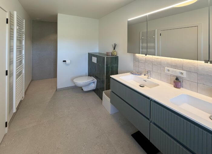 Salle de bains moderne équipée d'un meuble-lavabo suspendu, de deux vasques, d'un WC suspendu, d'un grand miroir avec éclairage LED, ainsi que d'un carrelage neutre au sol et aux murs. Un sèche-serviettes est fixé sur le mur de gauche. L'espace est minimaliste et épuré.
