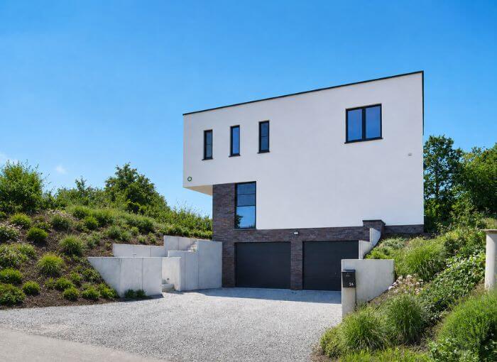 Maison moderne à deux étages, avec une façade blanche rehaussée de touches de briques foncées, de grandes fenêtres et deux portes de garage, nichée sur une pente herbeuse sous un ciel d'un bleu limpide.