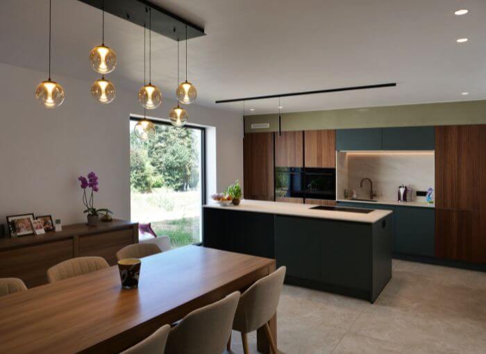 Cuisine et coin repas modernes, avec des placards en bois, des touches de vert, des suspensions, une grande fenêtre, des plantes d'intérieur et une table à manger en bois accompagnée de chaises beiges. La lumière naturelle illumine l'espace.